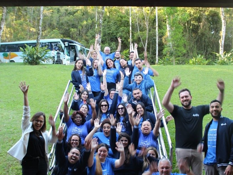 Turma de um encontro corporativo comemora ao ar livre com os braços erguidos, em escadaria cercada de mata