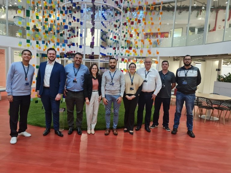 Grupo de profissionais posa lado a lado no átrio de uma empresa, sob uma instalação colorida suspensa