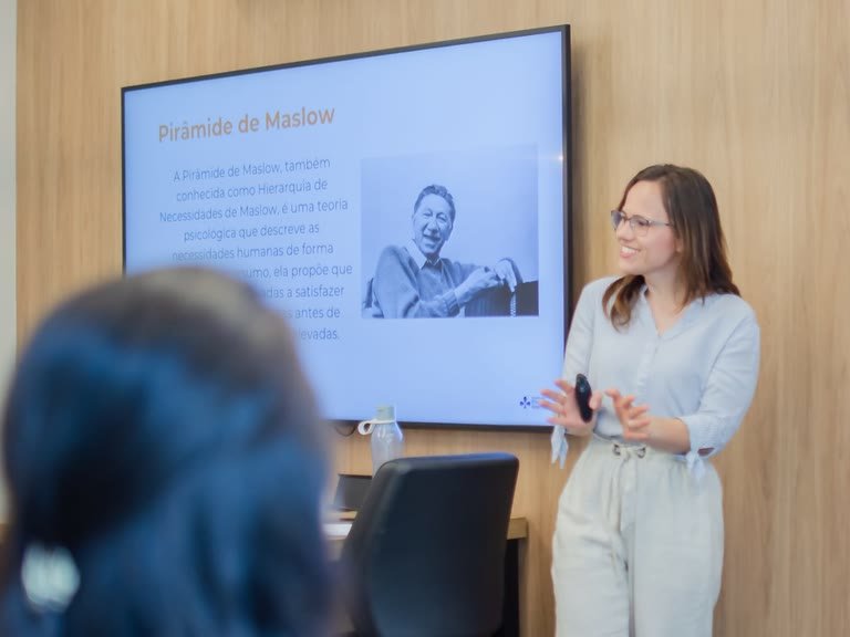 Palestrante em pé, gesticulando ao apresentar um slide sobre a Pirâmide de Maslow exibido na televisão da sala