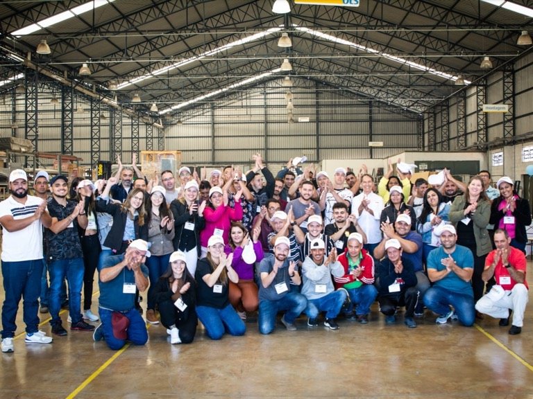 Turma reunida no galpão de uma indústria aplaude para a foto, em pé e agachada, muitos usando bonés do evento